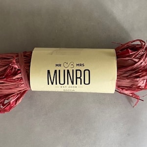 May include: A bundle of pink raffia with a cream-colored label wrapped around it. The label reads "MR & MRS MUNRO EST 2008 RAFFIA" in black text. The raffia is a vibrant shade of pink.