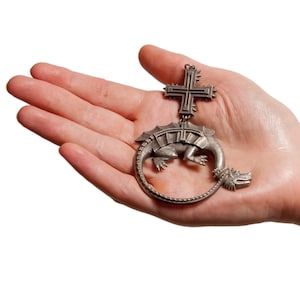 Puede incluir: Un colgante de plata con un dragón enroscado con una cruz en la espalda. El dragón está detallado con escamas y tiene una superficie texturizada.