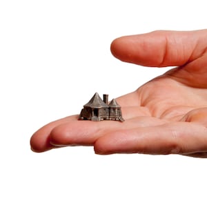 Puede incluir: Una casa en miniatura de color plateado con un diseño detallado, que incluye una chimenea y un techo puntiagudo. La pequeña casa se sostiene en la palma de una mano sobre un fondo blanco. La casa parece ser de metal.