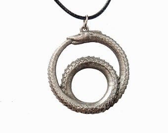 May include: A silver-toned metal pendant shaped like a serpent biting its own tail, forming a circular Ouroboros design with visible scale textures. The pendant hangs from a thin black cord attached by a small metal jump ring at the top.