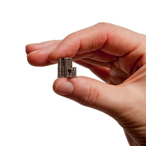 May include: A small, silver-coloured castle model featuring multiple towers and architectural details. The miniature castle is held between fingers against a white backdrop. The model appears to be made of metal.