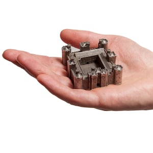 Op de afbeelding: Een klein, zilverkleurig metalen model van een kasteel met een vierkante basis en meerdere torens. Het model is gedetailleerd met ramen en deuren, en het dak is plat.
