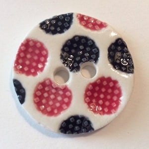 May include: A round, white button with two holes and a polka dot design. The button features red and navy blue polka dots with a textured pattern. This decorative button is suitable for sewing and craft projects.