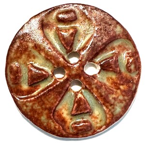 May include: A round, ceramic button with a cross-like design. The button features a textured, reddish-brown surface with a glossy finish. The design includes four triangular shapes and four holes for sewing. The button has a light green accent color.