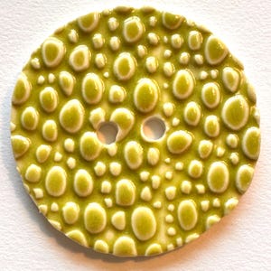 Könnte beinhalten: Ein runder Keramikknopf in hellem Grün mit strukturierter Oberfläche und erhabenen, ovalen Noppen. Er hat zwei Löcher und eine glänzende Oberfläche. Der Knopf ist vor einem weißen Hintergrund platziert.