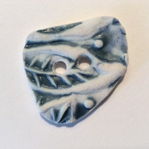 May include: A ceramic button with a leaf design. The button is a mix of white and blue, with two holes for sewing. The leaf pattern is carved into the surface, and the button has a slightly irregular shape.