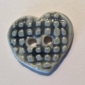 May include: A heart-shaped button with a textured surface and two holes. The button is a light gray color with a darker gray grid pattern. This decorative button is perfect for sewing projects.