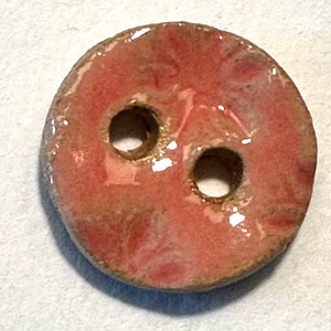 May include: A round, ceramic button with two holes. The button has a reddish-pink center with a marbled effect and a light brown edge. The button is glazed, giving it a glossy appearance.