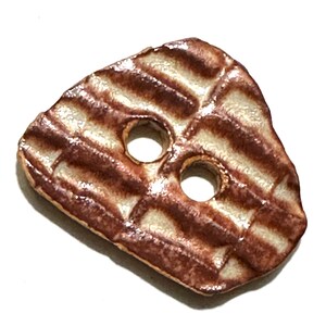 May include: A unique, ceramic button with a textured surface and two holes. The button features a brown and cream color scheme with a grid-like pattern. The button is irregularly shaped and appears handmade.