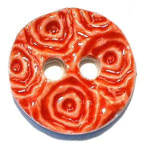 May include: A round, ceramic button with a glossy, red-orange finish. The button features a raised, swirling pattern and two evenly spaced holes for sewing. The design resembles a series of concentric circles.