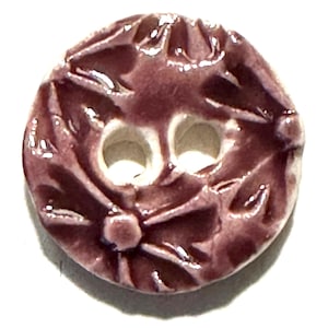 May include: A round, ceramic button with a glossy, dark purple glaze. The button features a floral design with two holes and a central raised dot. The button's design is embossed with leaf-like patterns.