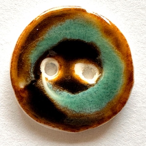 Può includere: Un bottone in ceramica fatto a mano con un design unico. Il bottone presenta un'area nera centrale circondata da un anello turchese e un bordo esterno marrone. Due fori bianchi sono visibili al centro. Il bottone è rotondo e lucido.