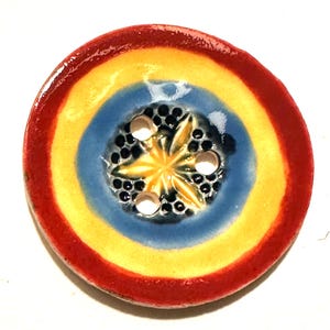 May include: A round, ceramic button with a vibrant design. The button features concentric rings of red, yellow, and blue. The center has a starburst design with small black dots and three white holes. The button is a decorative craft supply.