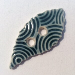May include: A ceramic button with a unique, irregular shape. The button is light blue with a dark blue wave pattern. It has two holes for sewing. The button's edges are uneven, giving it a handcrafted appearance.