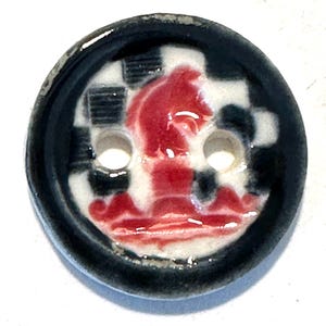 Puede incluir: Un botón de cerámica con borde negro y dos agujeros. La cara del botón presenta un caballo de ajedrez rojo sobre un fondo a cuadros blanco y negro. El botón es redondo y tiene un acabado brillante.