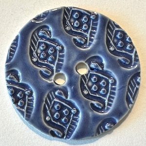 Könnte beinhalten: Ein runder, blauer Keramikknopf mit zwei Löchern. Der Knopf hat ein sich wiederholendes Muster aus weiß umrandeten Formen mit kleinen, erhabenen Punkten. Der Knopf hat eine glänzende Oberfläche.