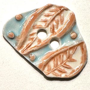 May include: A ceramic button with a light blue and cream color scheme. The button features two holes and is decorated with orange leaf patterns and small, round accents. The button has a glossy finish.