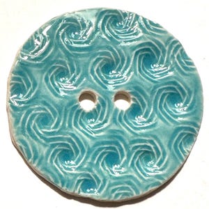 May include: A round, ceramic button with a glossy, turquoise glaze. The button features a repeating swirl pattern and two evenly spaced holes for sewing. The edges are slightly irregular, giving it a handmade appearance. The button is suitable for various sewing projects.