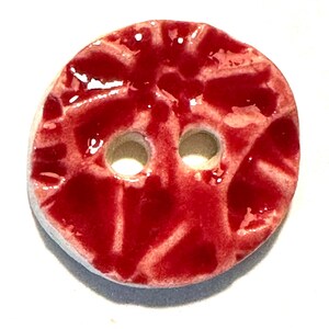 Op de afbeelding: Een ronde keramische knoop met twee gaten. De knoop heeft een glanzend, rood glazuur met een getextureerd, gekraakt patroon. De randen zijn licht, gebroken wit.