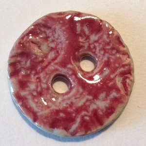 Può includere: Bottone rotondo in ceramica fatto a mano con due fori. Il bottone presenta un design marmorizzato lucido nei toni del rosso e del bianco. I bordi sono leggermente irregolari, conferendogli un aspetto rustico. Ideale per progetti di cucito.