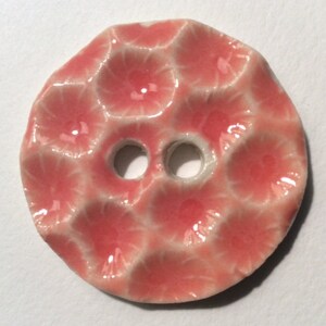 May include: A round, ceramic button with a textured surface and two holes. The button is a coral pink color with a glossy finish. The design features a pattern of raised, rounded shapes.