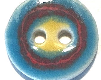 Puede incluir: Un botón redondo con dos agujeros, con círculos concéntricos de color. El anillo exterior es azul claro, seguido de un anillo rojo y un centro amarillo. El botón es probablemente de cerámica o vidrio.