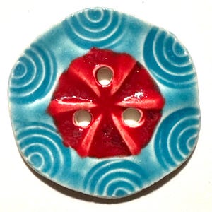 Op de afbeelding: Een keramische knoop met een rood bloemmotief in het midden en drie gaten. De knoop is omgeven door een lichtblauwe achtergrond met een spiraalpatroon. De knoop is rond en heeft een glanzende afwerking. Voor naaiwerk.