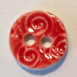 May include: A round, red ceramic button with three holes. The button has a glossy finish and a raised, swirling design. The edges are white. This button is suitable for sewing projects.