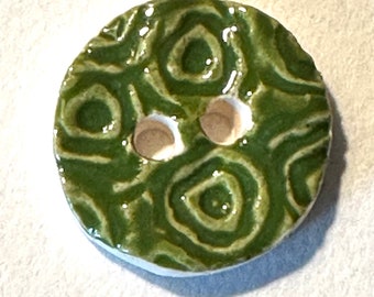 Puede incluir: Botón redondo de cerámica con un esmalte verde brillante y un diseño en espiral. El botón tiene dos agujeros y un borde blanco. El diseño recuerda a un motivo floral estilizado. Adecuado para proyectos de costura.