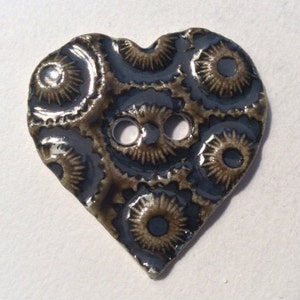 Pode incluir: Botão de cerâmica em forma de coração com design steampunk. O botão apresenta um fundo azul escuro com padrões castanhos em forma de engrenagens e dois orifícios para costura. O acabamento brilhante adiciona um aspeto refletor.