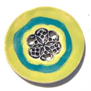 May include: A round ceramic button with a floral design in the center. The button features a yellow outer ring, a teal blue inner ring, and a dark gray floral pattern with small holes in the center.