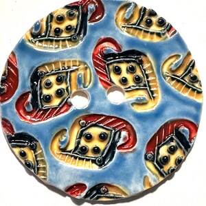 May include: A large, round button with a light blue background. The button features a repeating pattern of stylized, ornate figures in yellow, black, and red. The button has two holes for sewing.