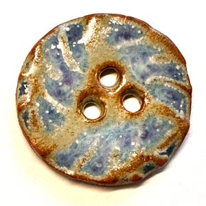 May include: A round ceramic button with three holes. The button features a swirling design in shades of blue, tan, and brown, with a speckled texture. The edges are a darker brown, giving it a rustic appearance.