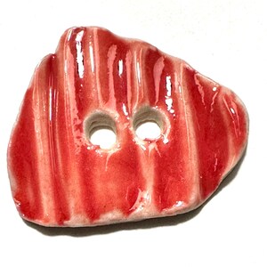 May include: A handmade ceramic button with a glossy red glaze. The button has a textured surface with vertical lines and two holes for sewing. The button is an irregular shape, resembling a piece of meat.