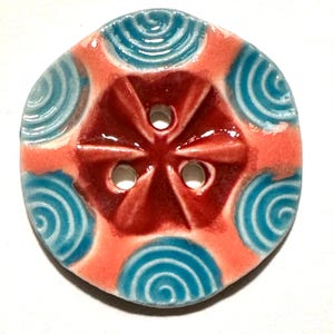 May include: A ceramic button with a circular shape. The button features a red center with three holes and a coral-colored outer ring. Teal spiral designs are evenly spaced around the edge, creating a decorative pattern. The button is likely for sewing or crafts.
