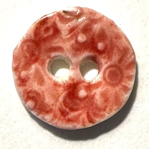 May include: A round, ceramic button with two holes. The button is primarily red with a marbled pattern and white accents. The button's design includes swirls and circular patterns.