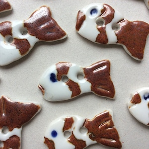 Puede incluir: Una colección de botones de cerámica con forma de pez. Cada botón presenta una base blanca con marcas marrones y blancas, una cola marrón y un punto azul para el ojo. Los botones tienen dos agujeros para coser.