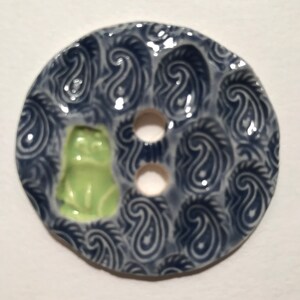 May include: A ceramic button with a navy blue paisley pattern and two holes. A small, light green frog is centered on the button. The button is round and has a glossy finish.