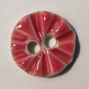 May include: A round, red button with a glossy finish. The button has a radial design with two holes for sewing. The button's design resembles a peppermint sweet.