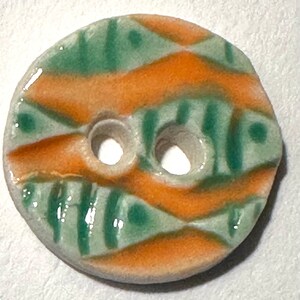 May include: A round ceramic button with two holes. The button features a pattern of orange and green diagonal stripes and shapes on a light gray background. The button is likely for sewing or crafts.
