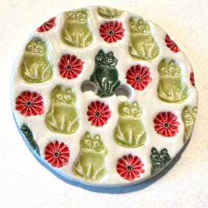 Puede incluir: Un jabonera redonda de cerámica blanca con un patrón repetido de figuras de gatos verdes y acentos de flores rojas. El centro presenta una figura de gato verde oscuro con dos agujeros. La jabonera tiene un acabado brillante.