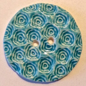 May include: A round, ceramic button with a glossy, turquoise glaze. The button features a repeating pattern of concentric circles, creating a textured surface. Two holes are visible for sewing. This decorative button is suitable for various sewing projects.