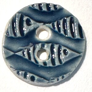 May include: A round, ceramic button with a glossy, dark blue glaze. The button features a textured design with two sets of parallel lines and two holes for sewing. The button's design is symmetrical and decorative.