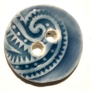 May include: A round, blue ceramic button with two holes. The button features a raised, swirling design with geometric and dotted patterns in a lighter blue color. The button is likely for sewing or crafts.