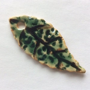 Puede incluir: Colgante de cerámica con forma de hoja con un agujero para colgar. La hoja es principalmente verde con vetas verde oscuro y un patrón moteado. Los bordes son de color marrón claro.