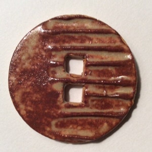May include: A round, ceramic button with a reddish-brown glaze and a textured, ribbed design. The button features two square cutouts and a lighter, tan-colored area. The button is likely handmade and suitable for sewing or crafts.