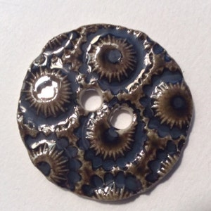 Pode incluir: Botão redondo de cerâmica com um design espiral azul e castanho. O botão tem três orifícios e um acabamento brilhante. O design apresenta padrões de espirais e estrelas repetidos, criando uma superfície texturizada. Adequado para projetos de costura e artesanato.