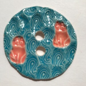 May include: A ceramic soap dish with a teal blue background and a swirling pattern. The dish features three holes and three small, orange cat figurines. The dish is round and has a slightly uneven edge.