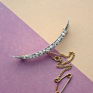 以下が含まれることがあります： 銀にセットされた透明な石の列が並んだ三日月型のブローチ。ブローチは安全ピン付きのゴールドチェーンに繋がっています。