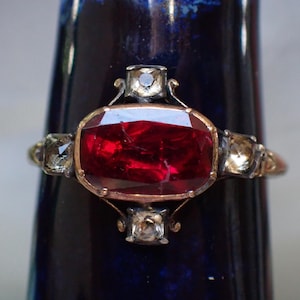 May include: A vintage ring with a large, red gemstone surrounded by smaller, clear gemstones. The ring is set in gold and has a delicate design.